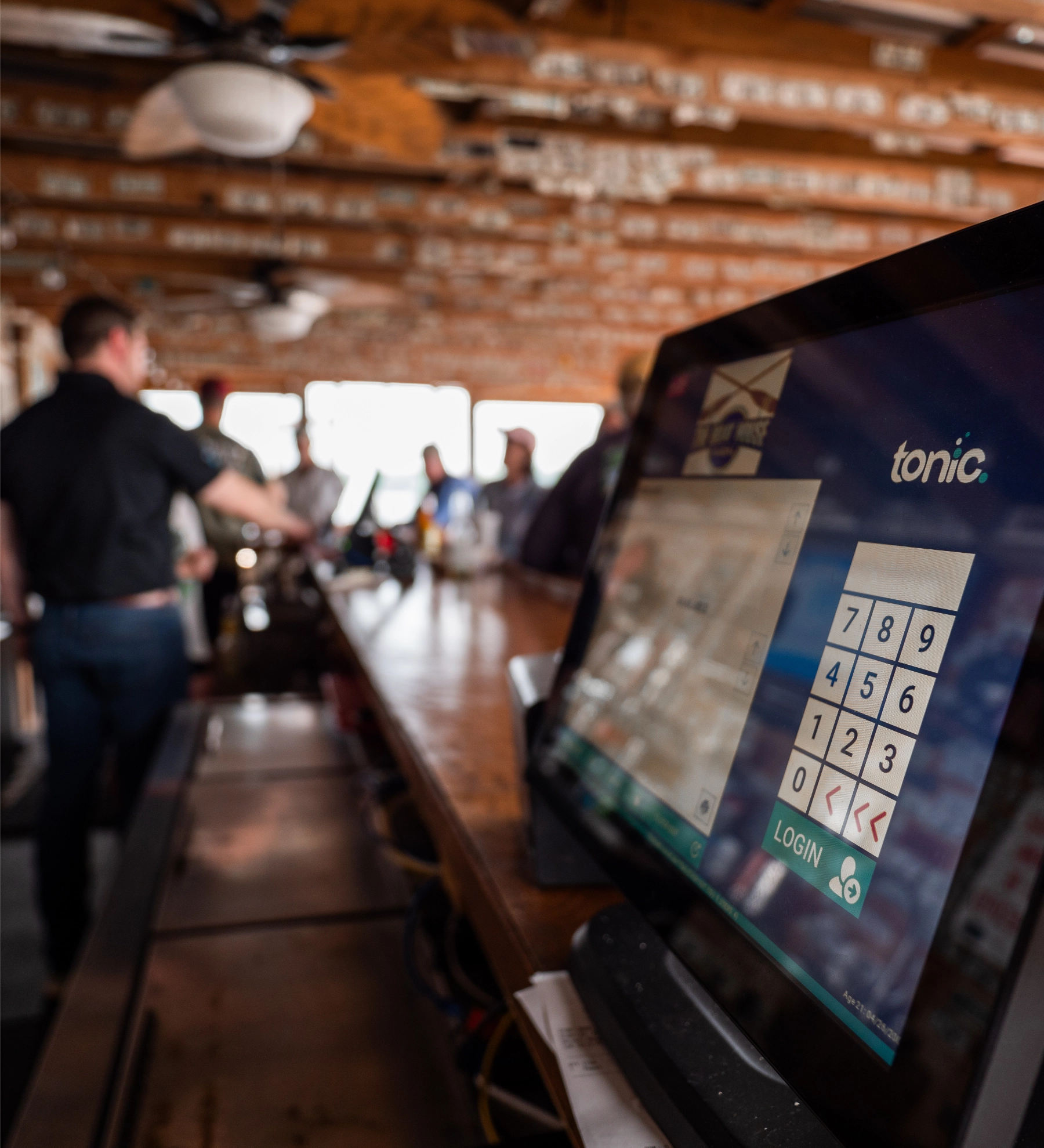 A Tonic POS countertop station sits on a restaurant bar counter, with blurred figures of a trainer and staff in the background.