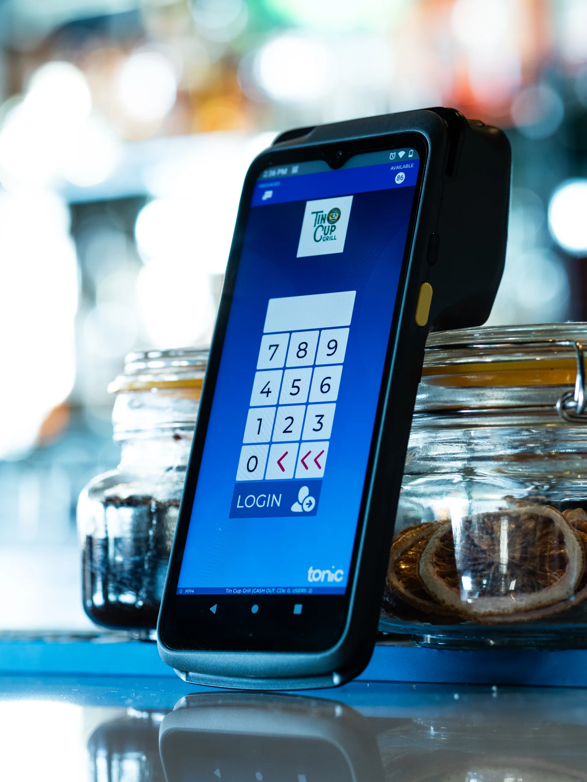 Tonic POS Handheld P8 - Upscale Bar Environment Dejavoo P8 handheld device with Tonic POS login screen, propped against cocktail jars on a high-end bar.